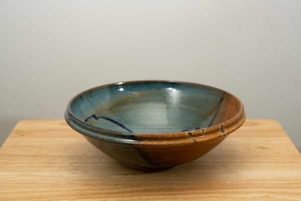 the photograph shows a clay coyote shallow salad bowl resting on a small light colored wooden table. the shallow salad bowl is glazed in joes blue, with the brown section of the glaze in the front right part of the bowl. the background is a white wall. the photograph is well lit with white light.