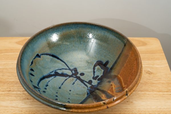 the photograph shows a high angle view of a clay coyote shallow salad bowl glazed in joes blue resting on a small light colored wooden table. the angle allows the viewer to see the details of the glaze pattern on the inside of the bowl. there is a section of brown, and darker blue splashes over the lighter blue base. the photograph is well lit with white light.