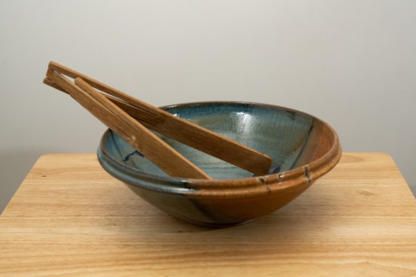 the photograph shows a clay coyote shallow salad bowl sitting on a small light colored wooden table. the shallow salad bowl is glazed in joes blue. the shallow salad bowl has a pair of baer woodworks foldable tongs resting inside the bowl. the tongs are resting on the left side of the bowl, and are made of a dark wood. the background is a white wall. the photograph is lit with white light.