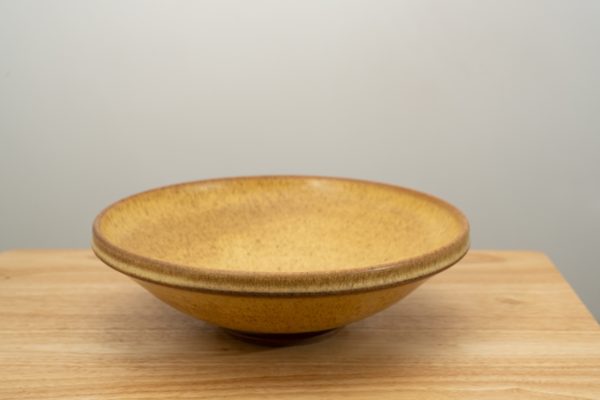the photograph shows a clay coyote shallow salad bowl glazed in yellow salt resting on a small light colored wooden table. the background is a white wall. the photograph is lit with white light.