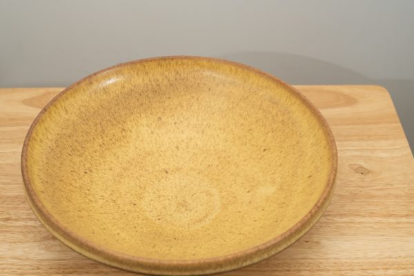the photograph shows a high angle view of a clay coyote shallow salad bowl glazed in yellow salt. the shallow salad bowl is resting on a small light colored wooden table. the background is a white wall. the photograph is well lit with white light