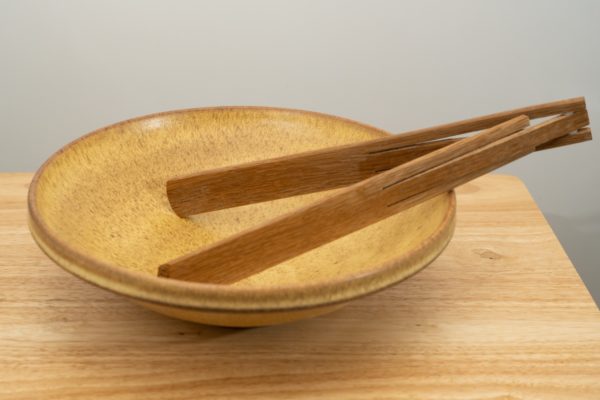 the photograph shows a clay coyote shallow salad bowl glazed in yellow salt sitting on a small light colored wooden table. the shallow salad bowl has a pair of baer woodworks foldable tongs resting in it. the tongs are resting on the right side of the bowl, and are made of dark wood. the background is a white wall. the photograph is lit by white light.