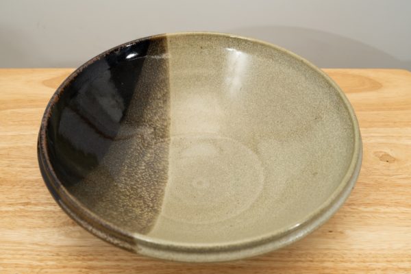the photograph shows a high angle view of a clay coyote shallow salad bowl glazed in mint chip resting on a small light colored wooden table. the angle is high enough to allow the viewer a good look at the glaze pattern on the inside of the bowl. there is a gradient of solid black into a mix of green and black finally into the mint green of the base glaze. the background is a white wall.