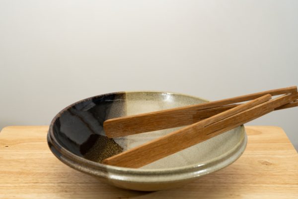the photograph shows a clay coyote shallow salad bowl glazed in mint chip sitting on a small light colored wooden table. the shallow salad bowl has a pair of baer woodworks foldable tongs resting in it. the tongs are in the open position, and are resting on the right side of the bowl. the tongs are made out of wood and are foldable. the background is a white wall. the photograph is well lit with white light.