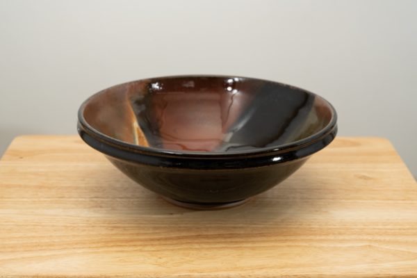 the photograph shows a clay coyote shallow salad bowl glazed in merlot resting on a small light colored wooden table. the background is a white wall. the photograph is lit with white light.