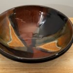 the photograph shows a high angle view of a clay coyote shallow salad bowl glazed in merlot. the angle of the photograph allows the viewer to see the detail of the glaze design on the inside of the bowl. the shallow salad bowl is resting on a small light colored wooden table. the background is a white wall. the photograph is well lit with white light.