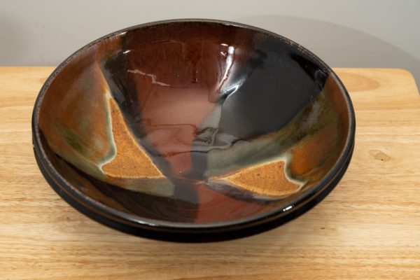the photograph shows a high angle view of a clay coyote shallow salad bowl glazed in merlot. the angle of the photograph allows the viewer to see the detail of the glaze design on the inside of the bowl. the shallow salad bowl is resting on a small light colored wooden table. the background is a white wall. the photograph is well lit with white light.