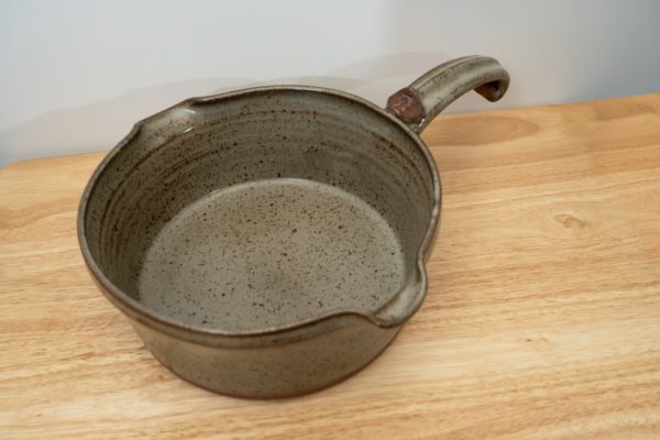 the photograph shows a clay coyote flameware medium saucepan resting on a small light colored wooden table. the flameware medium saucepan is resting flat on the table. the handle is pointed back and away from the viewpoint. the handle is pointed "towards" the upper right corner of the photograph. the background is a white wall. the photograph is lit with white light.