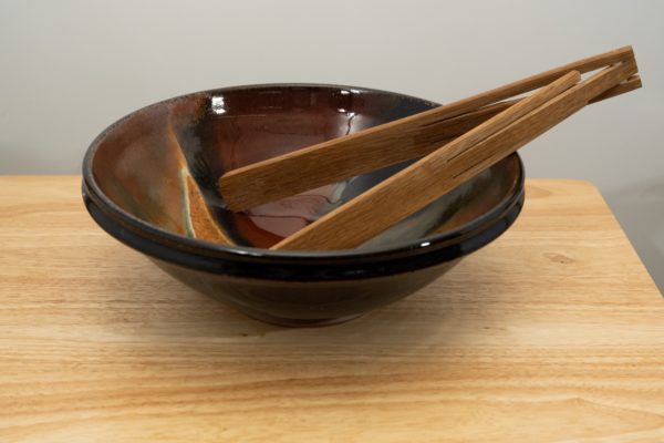 the photograph shows a clay coyote shallow salad bowl glazed in merlot sitting on a small light colored wooden table. the shallow salad bowl has a pair of baer woodworks foldable tongs resting in it. the tongs are in the open position, and are resting on the right side of the bowl. the tongs are made out of wood and are foldable. the background is a white wall. the photograph is well lit with white light.