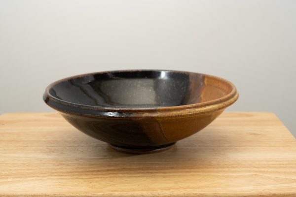 the photograph shows a clay coyote shallow salad bowl glazed in mocha swirl resting on a small light colored wooden table. the background is a white wall. the photograph is well lit with white light.