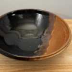 the photograph shows a high angle view of a clay coyote shallow salad bowl glazed in mocha swirl. the shallow salad bowl is resting on a small light colored wooden table. the angle of the photograph allows the viewer to see the inside of the bowl and the detail of the glaze pattern. the background is a white wall. the photograph is well lit with white light.