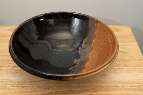 the photograph shows a high angle view of a clay coyote shallow salad bowl glazed in mocha swirl. the shallow salad bowl is resting on a small light colored wooden table. the angle of the photograph allows the viewer to see the inside of the bowl and the detail of the glaze pattern. the background is a white wall. the photograph is well lit with white light.