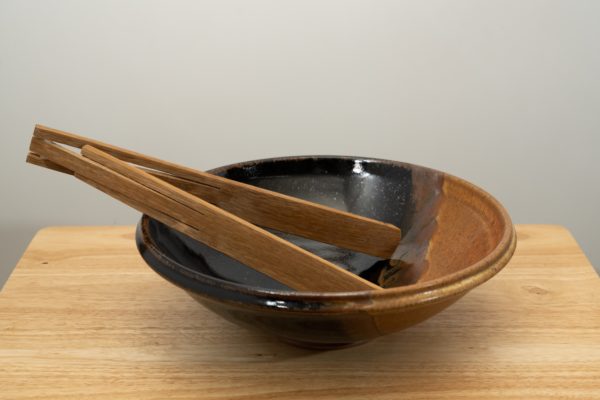 the photograph shows a clay coyote shallow salad bowl glazed in mocha swirl sitting on a small light colored wooden table. the shallow salad bowl has a pair of baer wooden tongs resting in it. the tongs are resting on the left side of the bowl. the background is a white wall. the photograph is well lit with white light.
