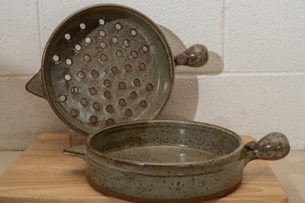 the picture shows the clay coyote inside-outside combination of grill basket and cazuela. the cazuela is resting flat on a small wooden table, with the main bulbous handle pointed to the right. the grill basket is behind the cazuela and is also on the small wooden table. the grill basket is resting on a clear plastic display piece allowing it to be vertical, to allow the viewer to see the grill baskets bottom, which has holes drilled into it. the grill baskets main bulbus handle is also pointed to the right. both the cazuela and the grill basket have secondary triangular handles on the opposite side of the pan from the bulbous handles. the triangle handle on both are pointed towards the left of the photograph. the background is a white painted cinderblock wall.