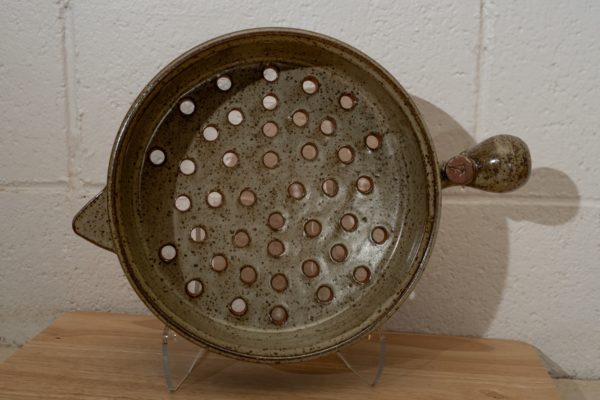 the photograph shows a clay coyote flameware grill basket resting on a small light colored wooden table. the grill basket is sitting in a clear plastic rest that allows it to be viewed vertically. this view shows off the bottom of the pan, showing the many holes in the grill baskets bottom. the primary bulbous handle is pointing towards the right, and the smaller secondary triangular handle is pointed towards the left. the background is a painted white cinderblock wall. the photograph was taken with a flash.