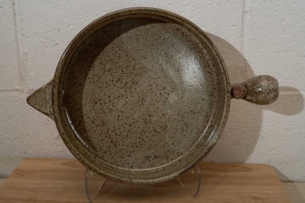 the photograph shows a clay coyote flameware cazuela resting on a light colored small wooden table. the cazuela is resting on a clear plastic display piece allowing it to be vertical. the cazuela is resting on its side allowing a clear view of the pans bottom. the small triangular handle is pointed to the left, and the large bulbous primary handle is pointed to the right of the photograph. the background is a painted white cinderblock wall.