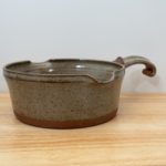 the photograph shows a clay coyote flameware medium saucepan resting on a small light colored wooden table. the flameware medium saucepan is resting flat on the table. the handle is pointed back and away from the viewpoint. the handle is pointed "towards" the upper right corner of the photograph. the background is a white wall. the photograph is lit with white light.