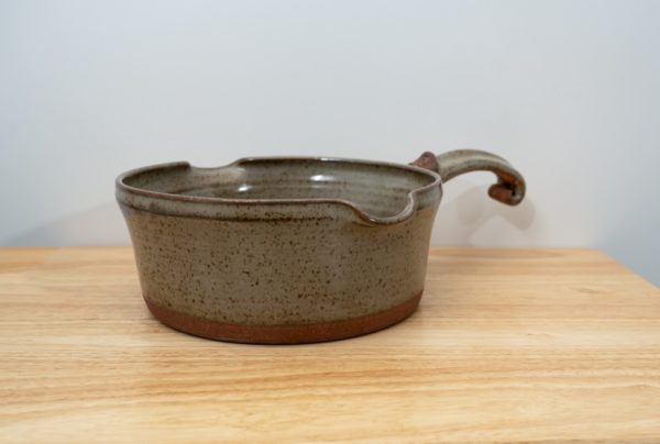 the photograph shows a clay coyote flameware medium saucepan resting on a small light colored wooden table. the flameware medium saucepan is resting flat on the table. the handle is pointed back and away from the viewpoint. the handle is pointed "towards" the upper right corner of the photograph. the background is a white wall. the photograph is lit with white light.