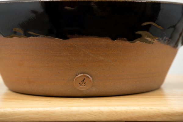 the photograph shows the bottom portion of a clay coyote special edition "Cassoulet Confessions" cassole resting on a light colored wooden surface. the cassole is glazed on the top lip (and inside but not visible) in midnight black. the outside base of the cassole is unglazed and shows off the natural color of the clay (reddish brown). the cassole has one small button on the front (facing towards the camera) with the clay coyote logo stamped into it. the button is in the unglazed portion, and is also unglazed. the photograph is lit with white light.