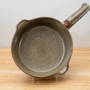 the photograph shows a clay coyote flameware medium saucepan resting on a small light colored wooden table. the flameware medium saucepan is resting on a small clear plastic stand, allowing the pan to be almost vertical. the handle for the pan is at the "2 o'clock" position on the near vertical pan. the pouring spouts on the pan are at the "11 o'clock" and "5 o'clock" position. the background is a white wall the photograph is lit with white light.