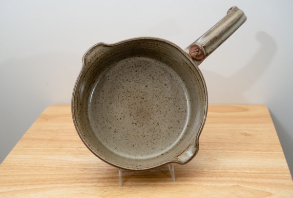 the photograph shows a clay coyote flameware medium saucepan resting on a small light colored wooden table. the flameware medium saucepan is resting on a small clear plastic stand, allowing the pan to be almost vertical. the handle for the pan is at the "2 o'clock" position on the near vertical pan. the pouring spouts on the pan are at the "11 o'clock" and "5 o'clock" position. the background is a white wall the photograph is lit with white light.