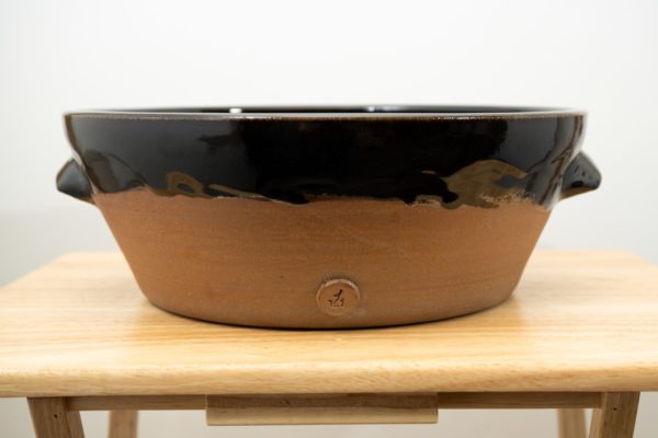 the photograph shows a full view of the front of a clay coyote special edition cassoulet confessions cassole. the inside and upper lip on the outside is glazed in midnight black. the glaze goes to the bottom of the two handles on the sides of the cassole. after that the cassole is unglazed and shows off the natural color of the clay (reddish brown). the cassole is resting on a small light colored wooden table. the background is a plain white wall.