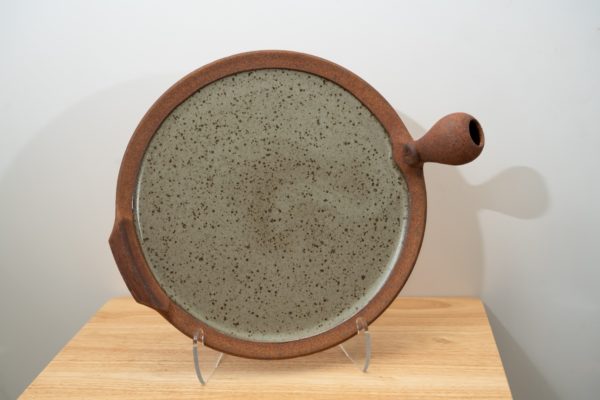 the photograph shows a clay coyote flameware pizza stone resting on a clear plastic stand. the stand allows the pizza stone to be viewed almost vertically. the viewpoint offered by this allows for the entire cooking surface to be seen. the bulbous handle is pointed to the right and the secondary triangular handle is pointed to the left. the pizza stones cooking surface is glazed in coyote grey (grey with dark speckling through out). the pizza stone is resting on a small light colored wooden table. the background is a white wall.