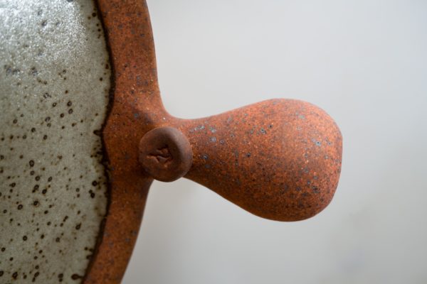 the photograph shows a clay coyote flameware pizza stone's main handle very close up. the photograph is taken close enough that only the bulbous main handle can be seen. the handle is pointed to the right. a small amount of the cooking surface can be seen on the left hand side of the photograph. the cooking surface is glazed in coyote grey (grey with dark speckling through out). the background is a plain white wall.