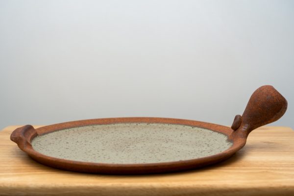 the photograph shows a clay coyote flameware pizza stone resting flat on a small light colored wooden table. the pizza stones bulbous handle is pointed towards the upper right corner of the photograph. the main body of the pizza stone is the cooking surface, and it is glazed in coyote grey (grey with dark speckling through out). the background is a white wall. the photograph is lit with white light.
