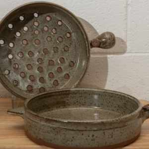 a horizontally framed photograph shows two clay coyote flameware cooking vessels. the clay coyote cazuela and the clay coyote grill basket. both of these pieces are glazed in coyote grey ( grey with darker speckling through out ). The grill basket is on a clear plastic stand, so it is standing on its side to allow full view of the grill basket holes in the bottom of the piece. the cazuela is sitting flat on the small wooden table. both the cazuela and the grill basket have their main handles pointed to the right side of the picture, and their smaller triangular secondary handles pointed to the left side. both pans are empty. behind them is a white painted cinder block wall.