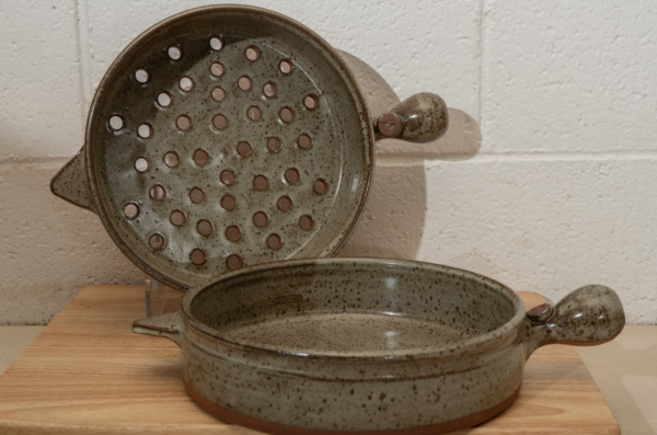 a horizontally framed photograph shows two clay coyote flameware cooking vessels. the clay coyote cazuela and the clay coyote grill basket. both of these pieces are glazed in coyote grey ( grey with darker speckling through out ). The grill basket is on a clear plastic stand, so it is standing on its side to allow full view of the grill basket holes in the bottom of the piece. the cazuela is sitting flat on the small wooden table. both the cazuela and the grill basket have their main handles pointed to the right side of the picture, and their smaller triangular secondary handles pointed to the left side. both pans are empty. behind them is a white painted cinder block wall.