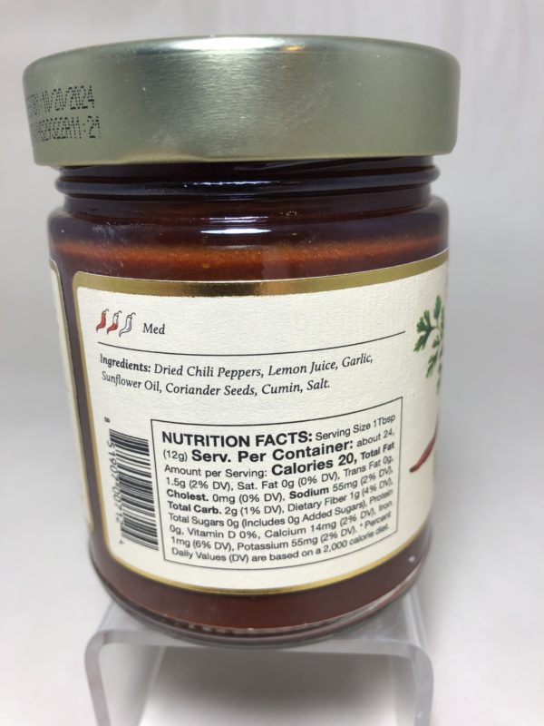 a close up horizontally framed photograph shows the ingredients ( dried chili peppers, lemon juice, garlic, sunflower oil coriander seeds, cumin and salt.) and nutrition facts. on the left side of the nutrition facts there is a UPC barcode. above the ingredient list there are three small peppers, with one and a half of them colored in red, and the word "Med" next to in indicating that the harissa is medium heat level.