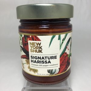 a horizontally framed photograph shows a close up view of the label on a bottle of New York Shuk brand signature harissa. the words "premium chili pepper condiment" are under the bold large letters NEW YORK SHUK and SIGNATURE HARISSA. The label has stylized chili peppers and its flowers on it. The Signature Harissa is sold Clay Coyotes website and in Gallery.