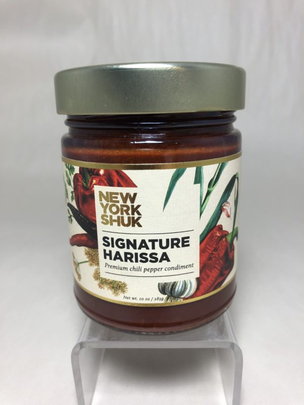 a horizontally framed photograph shows a close up view of the label on a bottle of New York Shuk brand signature harissa. the words "premium chili pepper condiment" are under the bold large letters NEW YORK SHUK and SIGNATURE HARISSA. The label has stylized chili peppers and its flowers on it. The Signature Harissa is sold Clay Coyotes website and in Gallery.