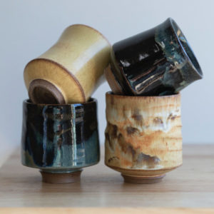 a square shaped photograph shows 4 yunomi wine cups sitting on a table, with a white background. the yunomis are sitting in two stacks of two. the left stack has a yellow salt glazed yunomi resting on top of a zappa glazed yunomi. the right stack has a midnight garden yunomi resting on top of a feather glazed yunomi.