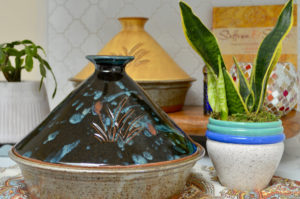a horizontally framed photograph shows two clay coyote tagines one in front of the other. the tagine in the back is glazed in yellow salt, the tagine in the front is glazed in midnight garden. to the right of the midnight garden (black base with a splash of blue and little bits of red and gold) is a snake plant in a white house plant pot. the house plant pot has three rings at the top of the pot. the lowest ring is blue, and the two rings on top of it are teal. directly behind the potted snake plant a small section of a dark blue colored pepper mill or shaker can be seen. also behind the snake plant, and to the right of the pepper mill is a reflective tiled candle holder or decorative bowl. the bowl has red, gold, and white tiles on it. behind the bowl and pepper mill is a yellow book with dark red writing on it. the word "saffron" and " & " can be made out on the cover, but everything else is out of focus. the book is leaning against the back wall behind the little table (the little table has the yellow salt tagine, the decorative bowl, pepper mill and book on it. the snake plant and the midnight garden tagine are on a separate table. this table has a white, soft blue red and orange paisley designed piece of cloth covering it. on the left side of the photograph partially out of frame is a house plant with green leaves and a small brown trunk visible. this houseplant is in a white ceramic pot with vertical lines going all the way around it. the room is well lit with white indoor lighting.