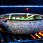 the photograph shows a clay coyote grill basket being used to cook mixed cut up vegetables on a lit grill. near the lower right corner of the photograph there is lit orange flame visible under the grills grate. the grill baskets handle is pointed to the left of the photograph, and is near by the lower left corner, just under halfway up the height of the photograph. the photograph was taken outside, so the photograph is lit with natural light.
