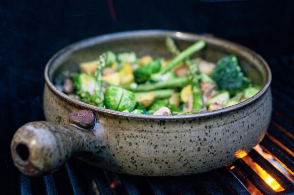 the photograph shows a clay coyote flameware grill basket being used to cook cut up mixed vegetables. the grill baskets handle is pointed almost directly towards the lower left hand corner of the photograph. the mixed vegetables in the grill basket are: brussel sprouts, broccoli, asparagus and potatoes. in the lower right corner of the photograph, orange flame flame is visible under the grill grate. the photograph was taken outside during the day.