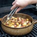the photograph shows a clay coyote flameware grill basket being used to cook diced potatoes on a grill. a hand is coming from the middle of the top edge of the photograph, reaching down with a pair of metal tongs to grab a few potatoes from the left side of the grill basket. there are yellow and red potatoes in the grill basket, with seasoning and crispy edges visible through out. the grill basket handle is pointed to the right of the photograph, with its smaller secondary triangular handle pointed to the left. the photograph was taken outside during the day.