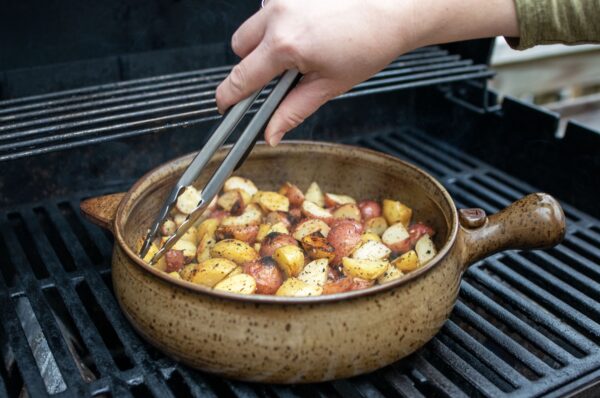 the photograph shows a clay coyote flameware grill basket being used to cook diced potatoes on a grill. a hand is coming from the middle of the top edge of the photograph, reaching down with a pair of metal tongs to grab a few potatoes from the left side of the grill basket. there are yellow and red potatoes in the grill basket, with seasoning and crispy edges visible through out. the grill basket handle is pointed to the right of the photograph, with its smaller secondary triangular handle pointed to the left. the photograph was taken outside during the day.