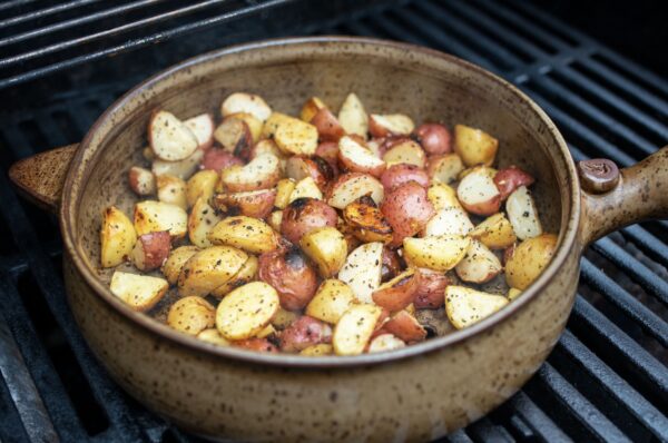 the photograph shows a clay coyote flameware grill basket being used to cook diced yellow and red potatoes on a grill. the potatoes have some crispy edges and there is seasoning visible through out. the handle of the grill basket is pointed towards the right side of the photograph. the photograph was taken outside during the day.