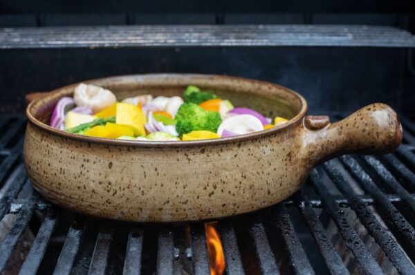 the photograph shows a clay coyote flameware grill basket being used on a lit grill to cook cut vegetables. there are yellow squash, broccoli, mushrooms, asparagus and purple onion. the grill baskets main handle is pointed to the right of the photograph. there is a small amount of orange flame visible under the grills grate, near the center of the lower edge of the photograph. the photograph was taken during the day.