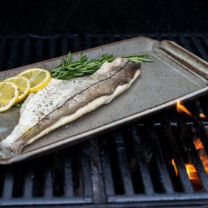 the photograph shows a clay coyote flameware fish tray being used on a lit grill. the fish tray has a fish fillet, 3 slices of lemon and a small bunch of herbs cooking on it. there is seasoning visible on the fish and lemon slices. in the bottom right corner of the photograph a small amount of orange flame is visible through the grill grates.
