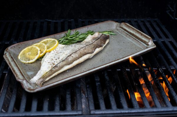 the photograph shows a clay coyote flameware fish tray being used on a lit grill. the fish tray has a fish fillet, 3 slices of lemon and a small bunch of herbs cooking on it. there is seasoning visible on the fish and lemon slices. in the bottom right corner of the photograph a small amount of orange flame is visible through the grill grates.