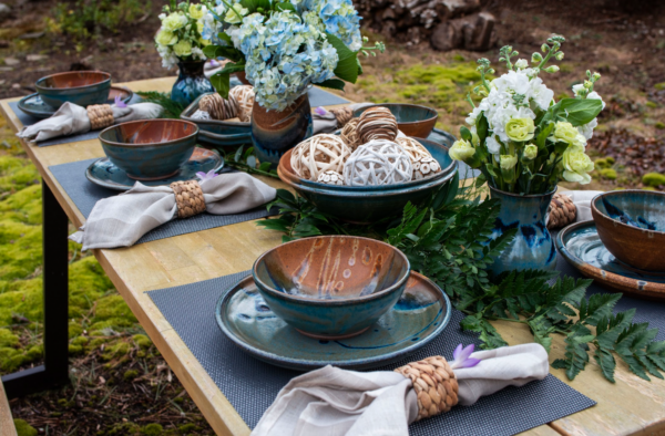 the photograph shows a long rectangular table with six clay coyote place settings around the table, three on each side. all the place settings are glazed in joes blue. there is a deep salad bowl glazed in joes blue also, it is near the center of the table. at the actual center of the table is a clay coyote large vase also glazed in joes blue. down the center of the table working from farthest away from the viewpoint to closest are these clay coyote pieces: a small vase, a baking dish, a large vase, deep salad bowl and finally a small vase. the vases all have flowers in them and the baking dish and deep salad bowl have decorative wooden balls in them. each place setting has a blue place mat, a dinner plate with a soup and chili bowl also glazed in joes blue on top of the dinner plate. there are linen napkins with what appears to be wicker napkin rings. each napkin/ring combo has a small purple flower tucked into the ring. the picture is outside, and under/behind the table there is visible moss. the photograph is lit with natural soft light.