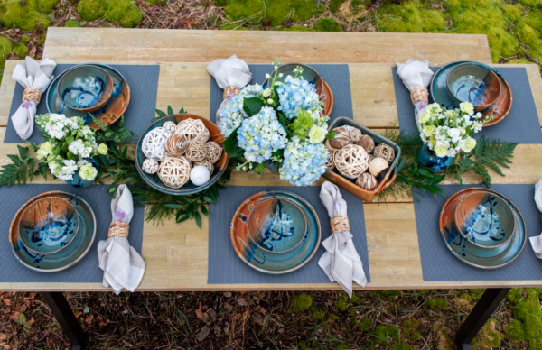 Joe's Blue Placesettings in Action the photograph shows a long rectangular table with 6 place settings on it. the table is longer left to right, then top to bottom. each one of the place settings has a blue placemat, then a clay coyote dinner plate with a clay coyote soup and chili bowl resting on top of it. to the right of each plate and bowl combo there is a linen napkin, with a wicker napkin ring. each napkin/ring set has a small purple flower tucked into the the ring. down the center of the table going left to right are the following: a clay coyote small vase with white flower bouquet, a clay coyote shallow salad bowl in joes blue filled with decorative balls, a large clay coyote vase in joes blue with a large blue bouquet in it, a clay coyote square baking dish also with decorative balls in it, and finally another small clay coyote vase. the table and everything on it is outside, and has clear white natural light.