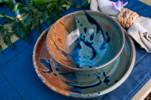 the photograph shows a clay coyote place setting close up. There are two clay coyote pieces in the photograph. there is a sandwich plate glazed in joes blue, with a soup and chili bowl also glazed in joes blue resting on it. the plate and bowl combo is resting on a dark blue placemat with a linen napkin next to it. the linen napkin is at the "2 o'clock" position relative to the bowl and plate. the linen napkin is off white or cream colored, it has a wicker style napkin ring on it. on the upper left corner of the photograph is a small branch of cut fern laying decoratively on the table. the photograph was taken outside, and has natural white light.