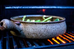 the photograph shows a clay coyote grill basket being used on a lit grill to cook cut up veggies. the cut up veggies include: asparagus, brussel sprouts, broccoli, yellow squash and mushrooms. there is little hints of orange glow from the flames below the grill basket. there are no visible flames, but there is orange light below the grill basket, and reflecting off the grill baskets main bulbous handle. the handle is pointed to the left of the photograph. the photograph is taken natural light.