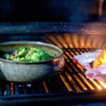 the photograph shows a clay coyote grill basket being used to grill cut up veggies on a lit grill. the grill basket takes up the left side of the photograph. the cut up veggies include asparagus, broccoli and other greens unidentifiable. on the right side of the grill and photograph there is two steaks being buttered on the grill. a spatula with a orange silicone head is being used to butter the steaks, it is coming from the lower right side. there are visible flames under the steaks, and visible blue flame under the grill basket.