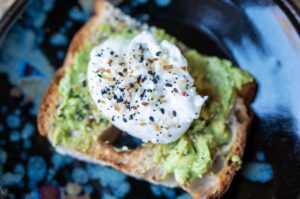 the photograph shows a clay coyote plate with zappa glaze being used to hold a piece of avocado toast, with a poached egg on top. both the avocado and poached egg have everything bagel seasoning sprinkled on them. the photograph is well lit.